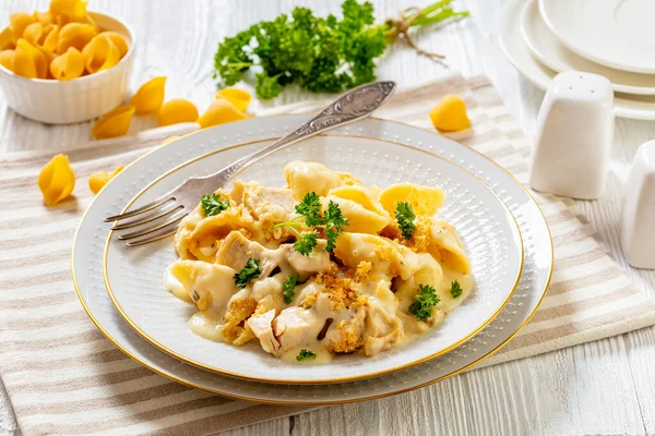 cheesy and creamy chicken and shells pasta bake in white bowl with fork on white table, horizontal view from above