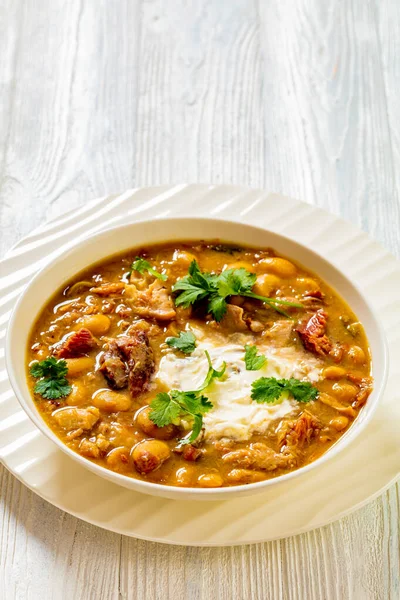 stock image smoked ham hock chicken chili in white bowl with fresh coriander and sour cream on white wood table, vertical view from above