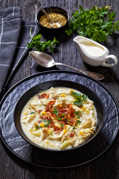 Chicken Corn rich and creamy soup with chicken breast, potatoes, sweet corn, topped with fried crispy bacon and fresh parsley in black bowl on wooden table, vertical view