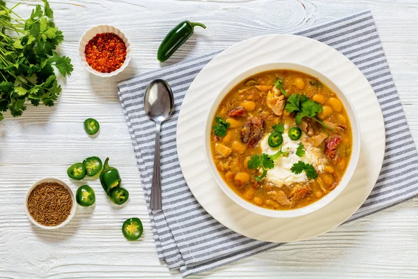 stock image smoked ham hock chicken chili in white bowl with fresh coriander and sour cream on white wood table, horizontal view from above, flat lay