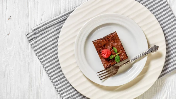 chocolate brownie with fresh raspberry and mint on top on a white plate with dessert fork, horizontal view from above, flat lay, free space