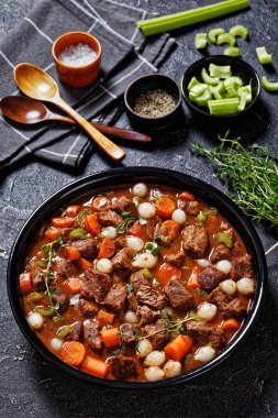 boneless beef short ribs stew with carrots and pearl onions in black bowl on concrete table with napkin and wood spoons, vertical view from above clipart