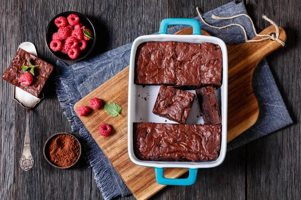 chocolate brownies with fresh raspberry and mint in a blue baking dish on cutting board on dark wood table, horizontal view from above, flat lay