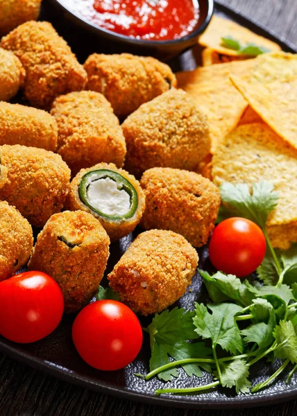 stock image close-up of breaded crispy fried jalapeno poppers with melted cream cheese filling with taco chips, tomatoes and salsa on black platter, macro