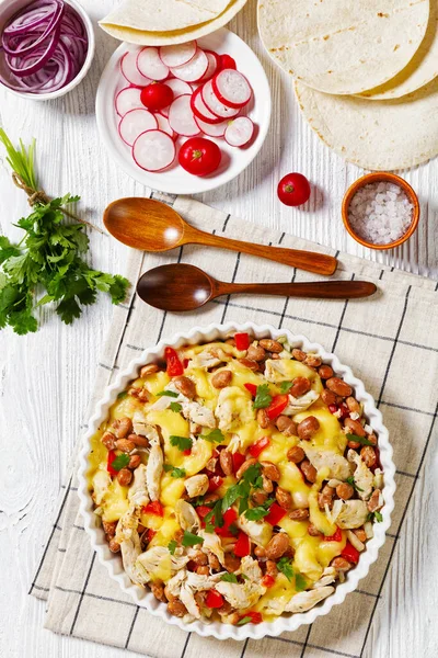 shredded chicken breast, pinto beans, tomato and mozzarella in white round baking dish on white wood table with wooden spoons, flatbreads, red radish, vertical view from above