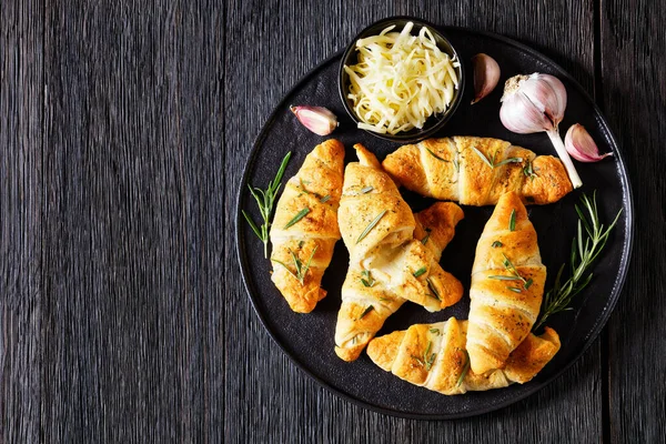 Stock image savory crescent rolls with melted cheese filling, sprinkled with fresh rosemary leaves on black plate on dark wooden table with garlic butter, horizontal view from above, flat lay, copy space