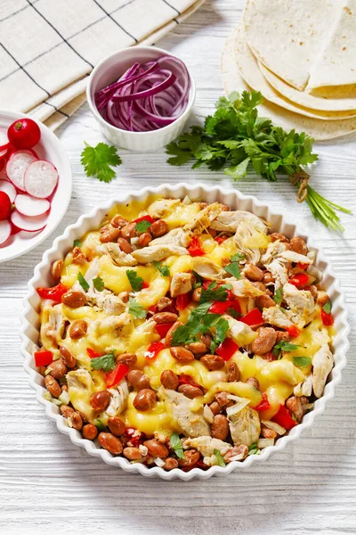 stock image shredded chicken breast, pinto beans, tomato and mozzarella in white round baking dish on white wood table with flatbreads, red radish, vertical view from above, close-up