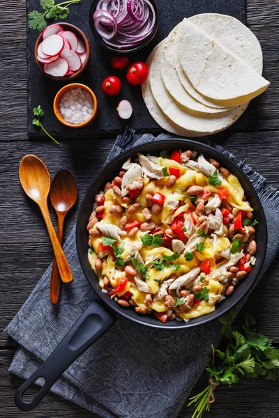 shredded chicken breast, pinto beans, tomato and mozzarella skillet on a dark wood table with wooden spoons, flatbreads, red radish, verticall view from above