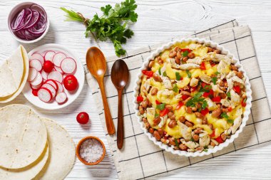 shredded chicken breast, pinto beans, tomato and mozzarella in white round baking dish on white wood table with wooden spoons, flatbreads, red radish, horizontal view from above, flat lay clipart