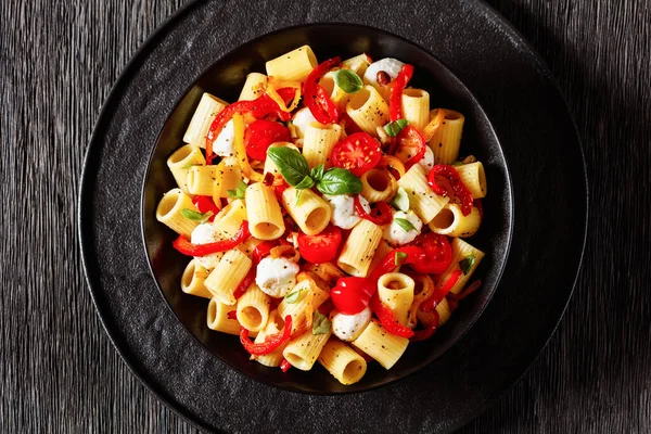 roasted pepper tomato mozzarella pasta salad in black bowl on dark oak wood table, horizontal view from above, flat lay, close-up
