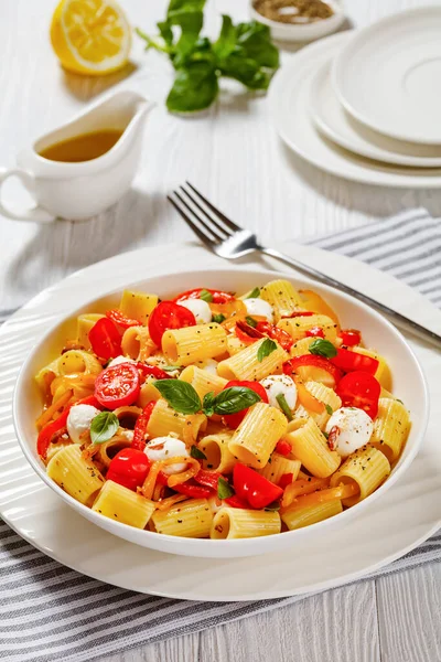 roasted pepper tomato mozzarella pasta salad in white bowl on white wood table, vertical view from above