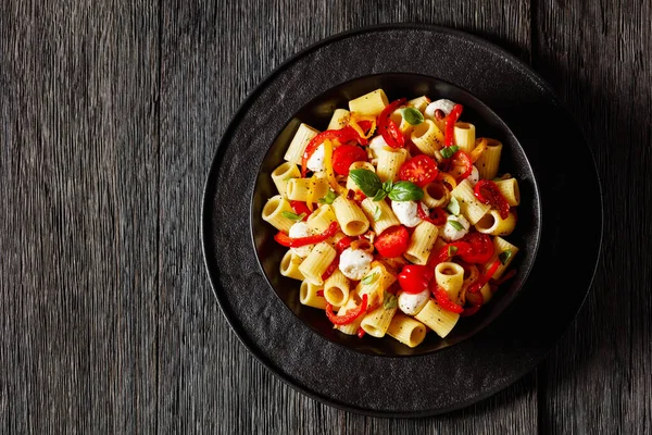 roasted pepper tomato mozzarella pasta salad in black bowl on dark oak wood table, horizontal view from above, flat lay, free space