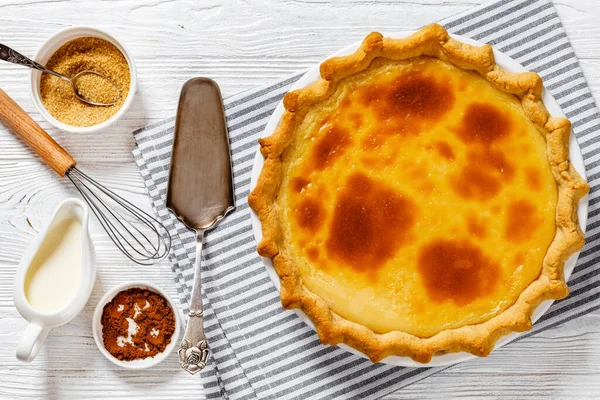 classic sugar cream pie, hoosier pie, indiana custard cream pie on white baking dish on white wood table with sugar, whisk, cinnamon, cake shovel, horizontal view from above, flat lay, close-up