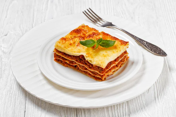 stock image portion of lasagne al forno, italian beef lasagna layered with ground beef, marinara sauce, pasta noodles and ricotta cheese on white plate with fork on white wood table