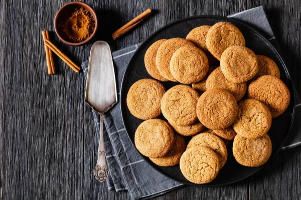 soft ginger cookies on black plate on dark oak table with cinnamon and cake shovel, horizontal view from above, flat lay, copy space
