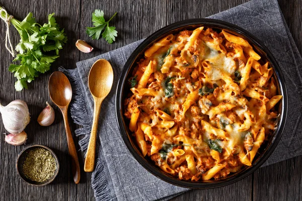 Ground Chicken Pasta Bake with onion, mushrooms, spinach, tomato sauce and mozzarella cheese in baking dish on dark wood table, horizontal view from above, flat lay