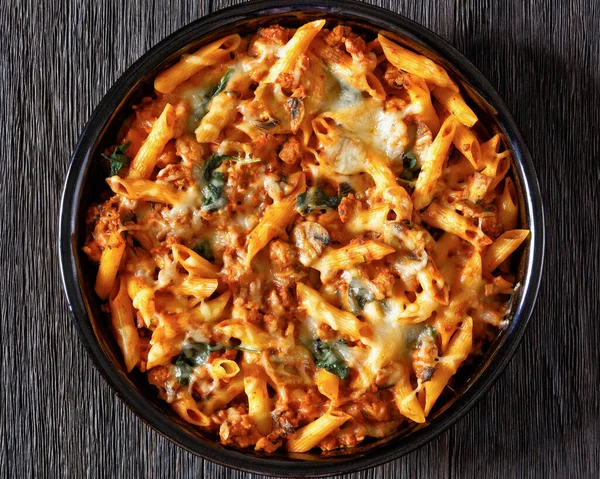 Ground Chicken Pasta Bake with onion, mushrooms, spinach, tomato sauce and mozzarella cheese in baking dish on dark wood table, horizontal view from above, flat lay, close-up