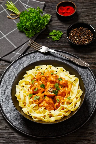 Stock image Creamy Paprika Pork Chunks over tagliatelle noodle in black bowl on dark oak wooden table, vertical view