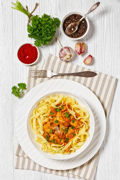 stock image Creamy Paprika Pork Chunks over tagliatelle noodle in white bowl on white wood table with ingredients, fork and napkin, vertical view from above