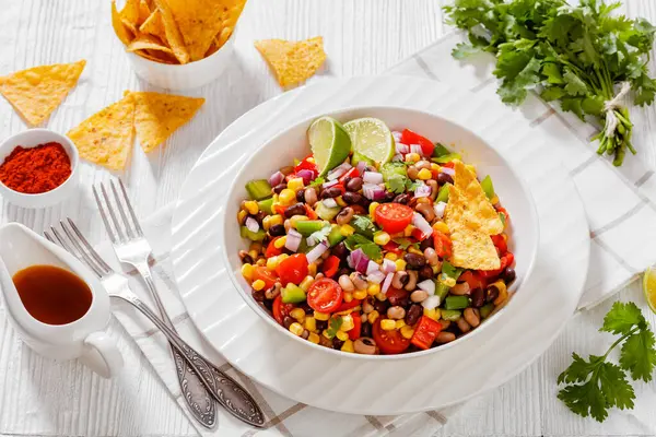 Black Bean Salad with Black-Eyed Peas, pepper, red onions, corn, cherry tomatoes and cilantro in white bowl with tortilla chips, nachos and vinaigrette dressing on wood table, landscape view, close-up