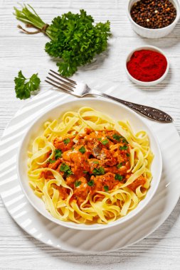 Creamy Paprika Pork Chunks over tagliatelle pasta in white bowl on white wood table with ingredients, fork and napkin, vertical view from above clipart
