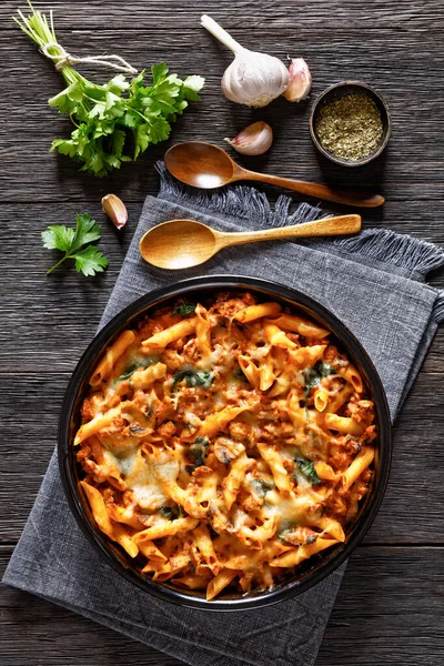 Ground Chicken Pasta Bake with onion, mushrooms, spinach, tomato sauce and mozzarella cheese in baking dish on dark wood table, vertical view from above