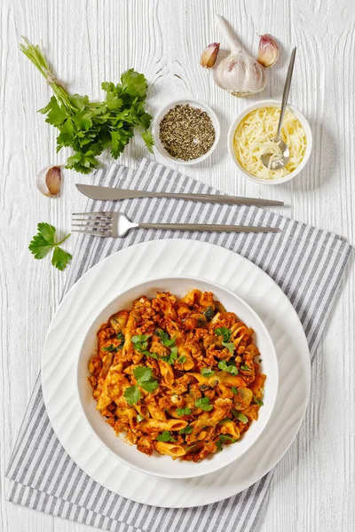 Ground Chicken Pasta Bake with onion, mushrooms, spinach, tomato sauce and mozzarella cheese in white bowl on white wood table, vertical view from above