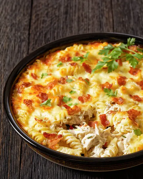 fusilli pasta bacon cheese chicken bake in baking dish on dark wooden table, vertical view from above, close-up