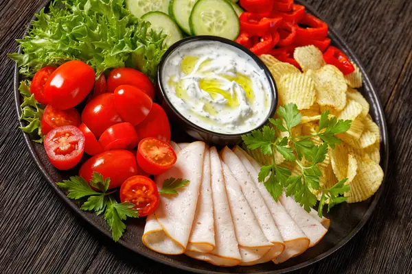 sliced ham, potato chips, leafy greens and vegetables snack platter with healthy yogurt ranch dip on dark wooden table, dutch angle view