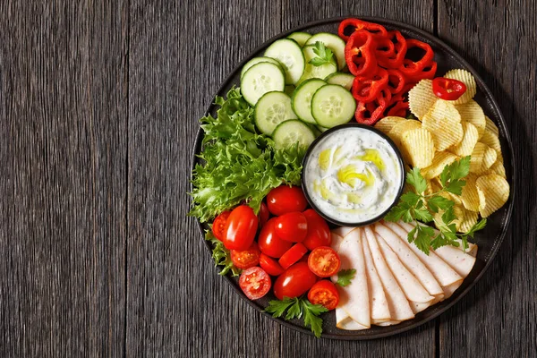 sliced ham, potato chips, leafy greens and vegetables snack platter with healthy yogurt ranch dip on dark wooden table, horizontal view from above, flat lay, free space