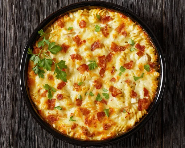 fusilli pasta bacon cheese chicken bake sprinkle with fresh parsley in baking dish on dark wooden table horizontal view from above, flat lay, close-up