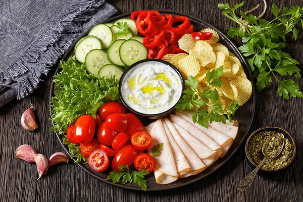 close-up of sliced ham, potato chips, leafy greens and vegetables snack platter with healthy yogurt ranch dip on dark wooden table