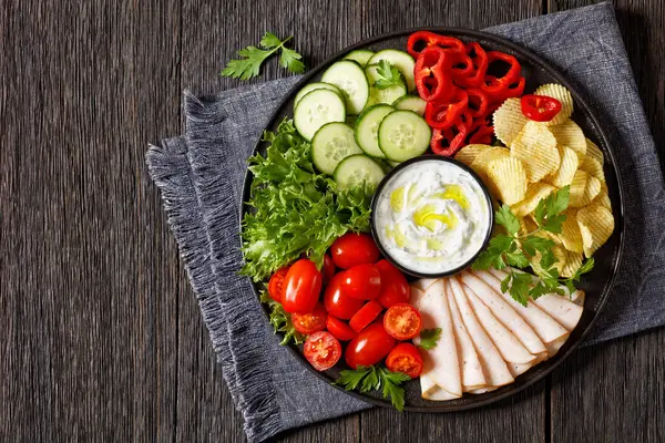sliced ham, potato chips, leafy greens and vegetables snack platter with healthy yogurt ranch dip on dark wooden table, horizontal view from above, flat lay, copy space