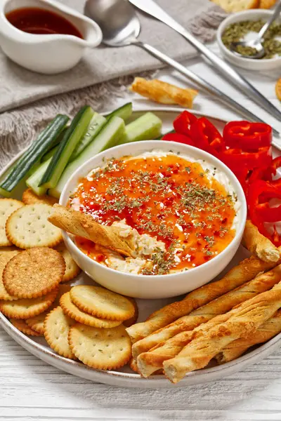 Hot Pepper Jelly Cheese Dip in bowl served with crackers, salty grissini, fresh pepper and cucumber on platter on white wooden table, vertical view, close-up