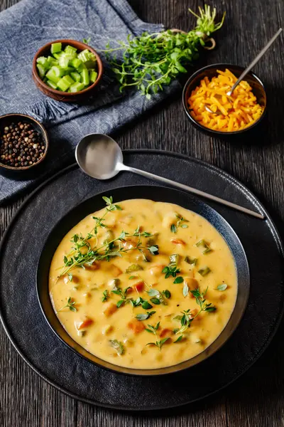 Beer cheese soup with vegetables and thyme in black bowl on dark wooden table with ingredients, spoon and tea towel, vertical view from above
