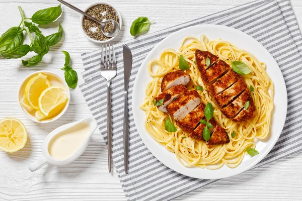 Chicken Scallopini of crusted chicken breast over lemon butter pasta on white plate on white wooden table, horizontal view from above, flat lay