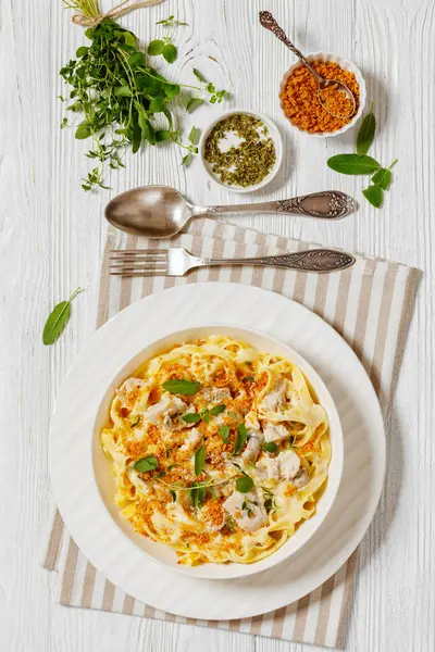 creamy chicken and mushroom tagliatelle topped with breadcrumbs and fresh herbs in white bowl on white wooden table, vertical view from above