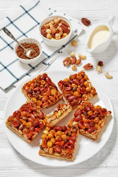 homemade toffee mixed nuts shortbread bars on white plate on white wooden table with ingredients, vertical view