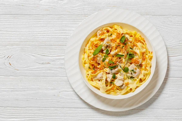 creamy chicken and mushroom tagliatelle topped with breadcrumbs and fresh herbs in white bowl on white wooden table, horizontal view from above, flat lay, free space