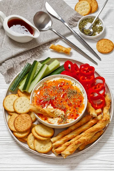 Hot Pepper Jelly Cheese Dip in bowl served with crackers, salty grissini, fresh pepper and cucumber on platter on white wooden table, vertical view