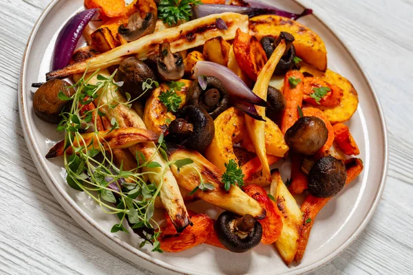 baked winter vegetables and mushrooms, potato, winter squash, red onions, carrots, parsnip on plate on white wooden table, dutch angle view, close-up