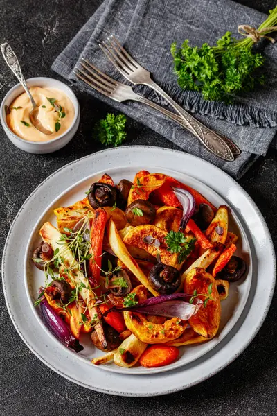 baked winter vegetables and mushrooms,  potato, winter squash, red onions, carrots, parsnip on plate on concrete table with forks and sauce, vertical view from above