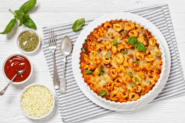 tortellini baked with mozzarella cheese, tomato sauce and ground beef in white baking dish on white wooden table, horizontal view from above, flat lay