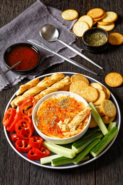 Hot Pepper Jelly Cheese Dip in bowl served with crackers, salty grissini, fresh pepper and cucumber on platter on dark wooden table, vertical view