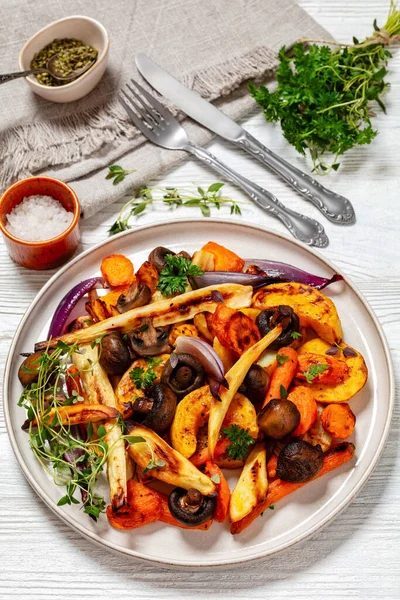 baked winter vegetables and mushrooms, potato, winter squash, red onions, carrots, parsnip on plate on white wooden table with cutlery and fresh herbs, vertical view