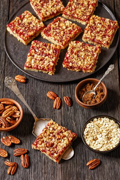 Red berry Shortbread Bars with Rolled Oat and Nut Topping on black plate on dark wooden table with ingredients and cake shovel, vertical view from above