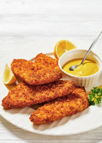breaded fish fillet baked in oven served with yellow mustard on white plate on white wooden table, vertical view, close-up