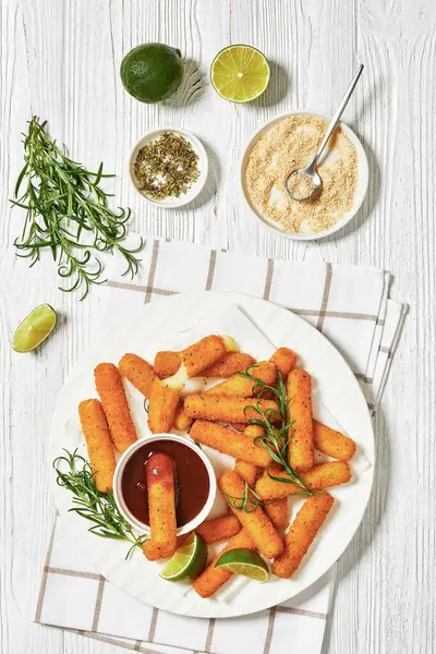 fried breaded mozzarella sticks served with dipping sauce, lime slices and fresh rosemary on white platter on white wooden table with ingredients, vertical view from above