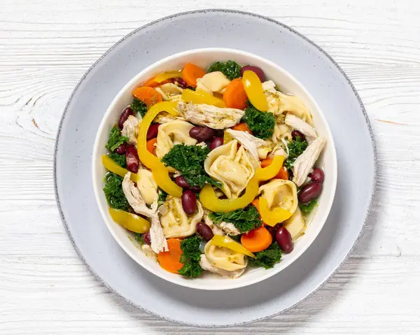 chicken minestrone soup with tortelloni and red kidney beans, kale and yellow bell pepper in bowl on white wooden table, horizontal view from above, flat lay, close-up
