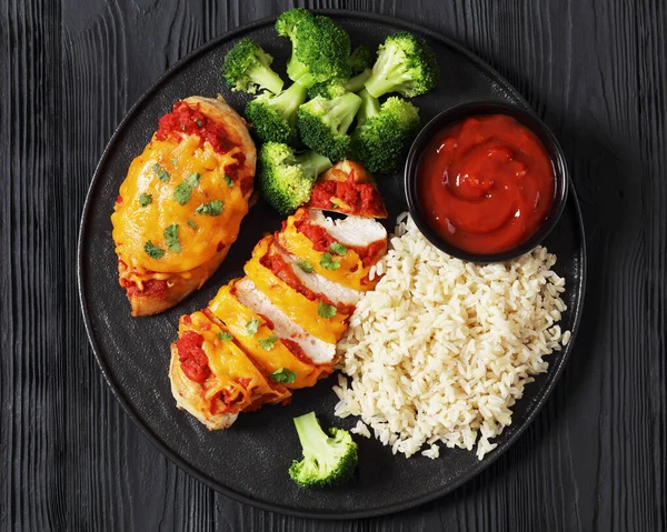 baked chicken breasts with tomato salsa, cheddar cheese and parsley on black plate with broccoli, brown rice and tomato sauce on black wooden table, horizontal view from above, flat lay, close-up